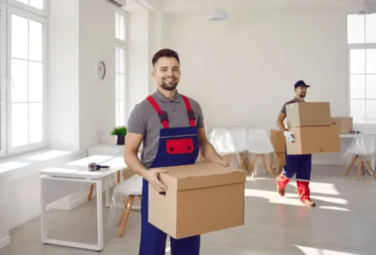 Un traslocatore che solleva una scatola, mentre un suo collega sullo sfondo ne sposta altre due