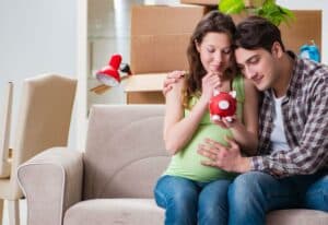 Donna in dolce attesa con salvadanaio rosso in mano e uomo che le tocca la pancia con scatoloni sullo sfondo